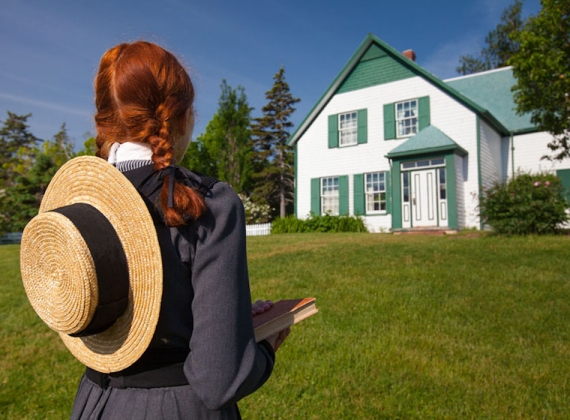A girl with red hair in braids and a sun hat on her back looking at a white house on a sunny day.